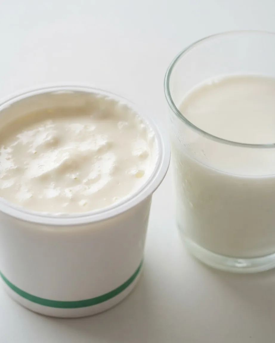 An open cup of thick white yogurt beside a clear glass of milk on a pale tabletop.