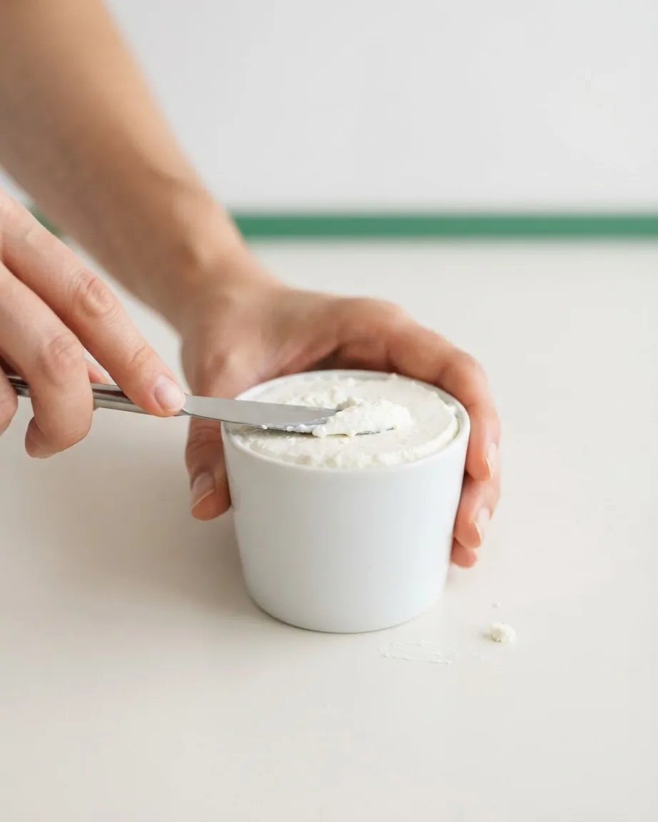 Hands level soft white ricotta at the rim of a small white cup with a metal knife.