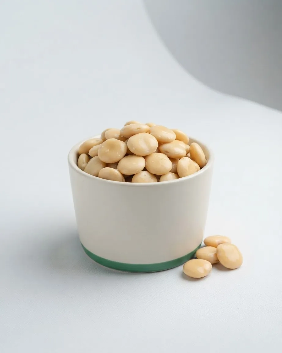 A white ceramic cup filled with pale yellow lupini beans, with a few beans scattered beside it on a neutral surface.