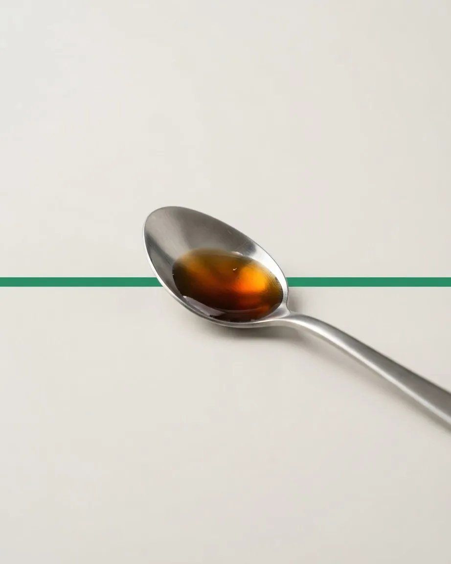 A stainless steel teaspoon holding amber-brown liquid rests on a pale surface crossed by a green horizontal line.