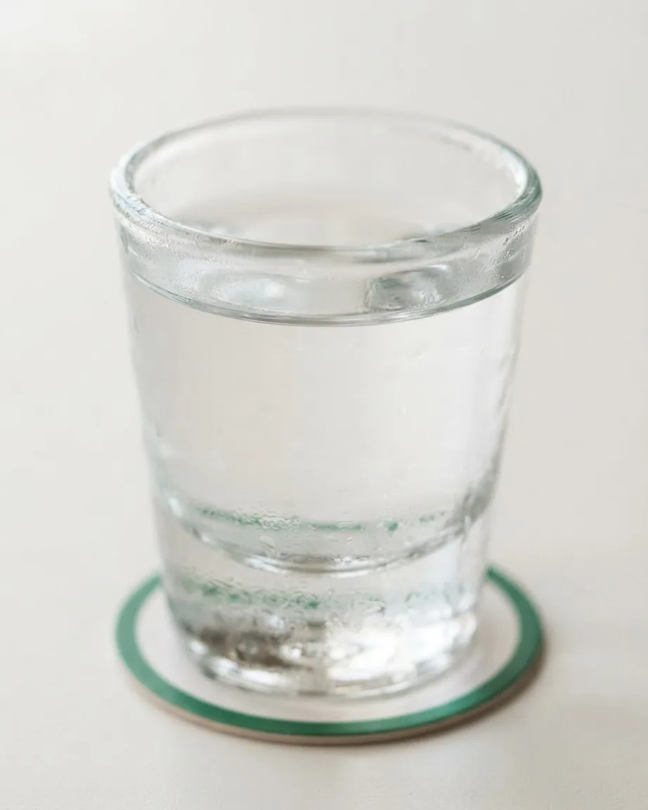 A clear shot glass filled with transparent liquid sits on a white coaster with a green rim against a pale background.